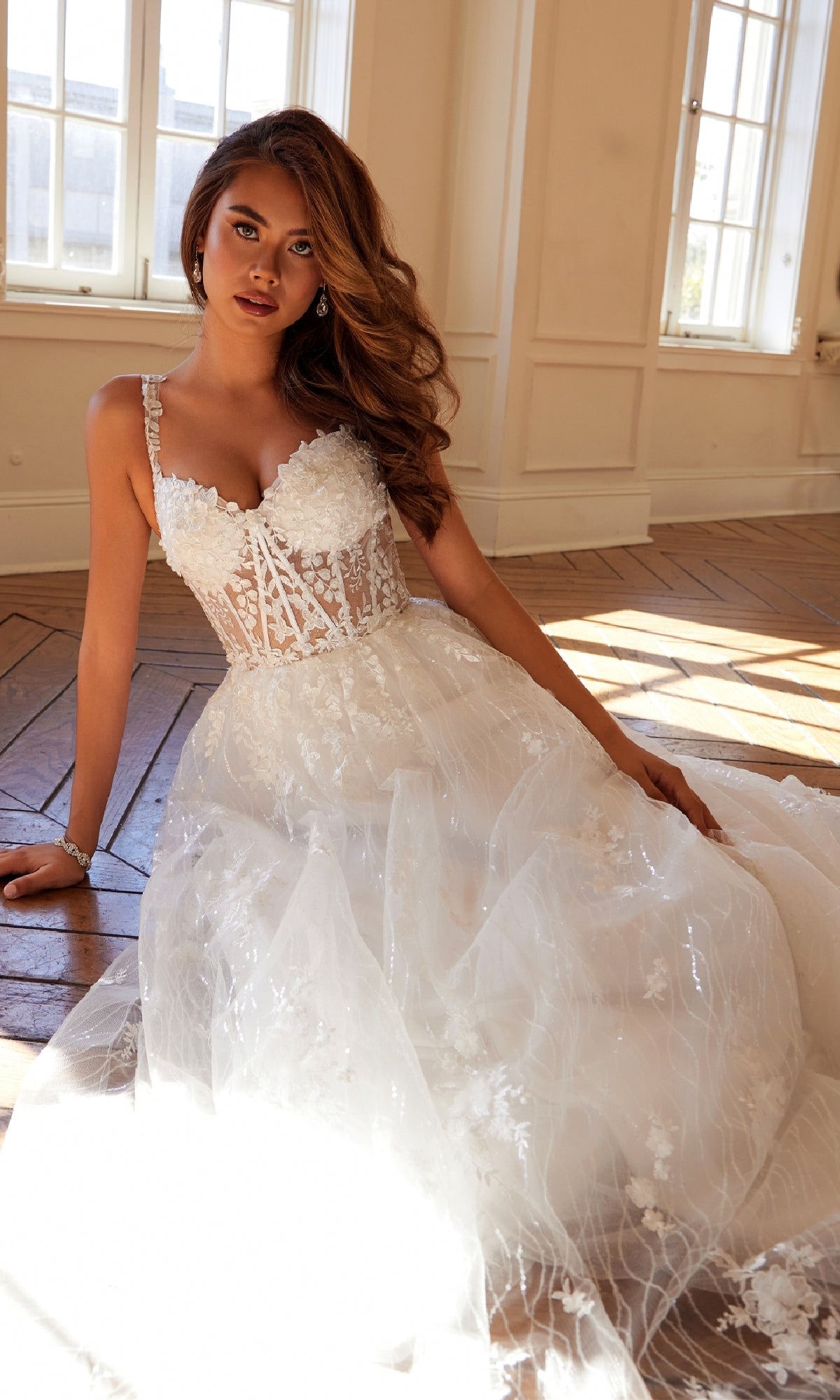 A woman wearing the Long Prom Dress: Juliet JT2495AW with a glittery ball gown and lace corset bodice sits on a wooden floor in a bright room with large windows.